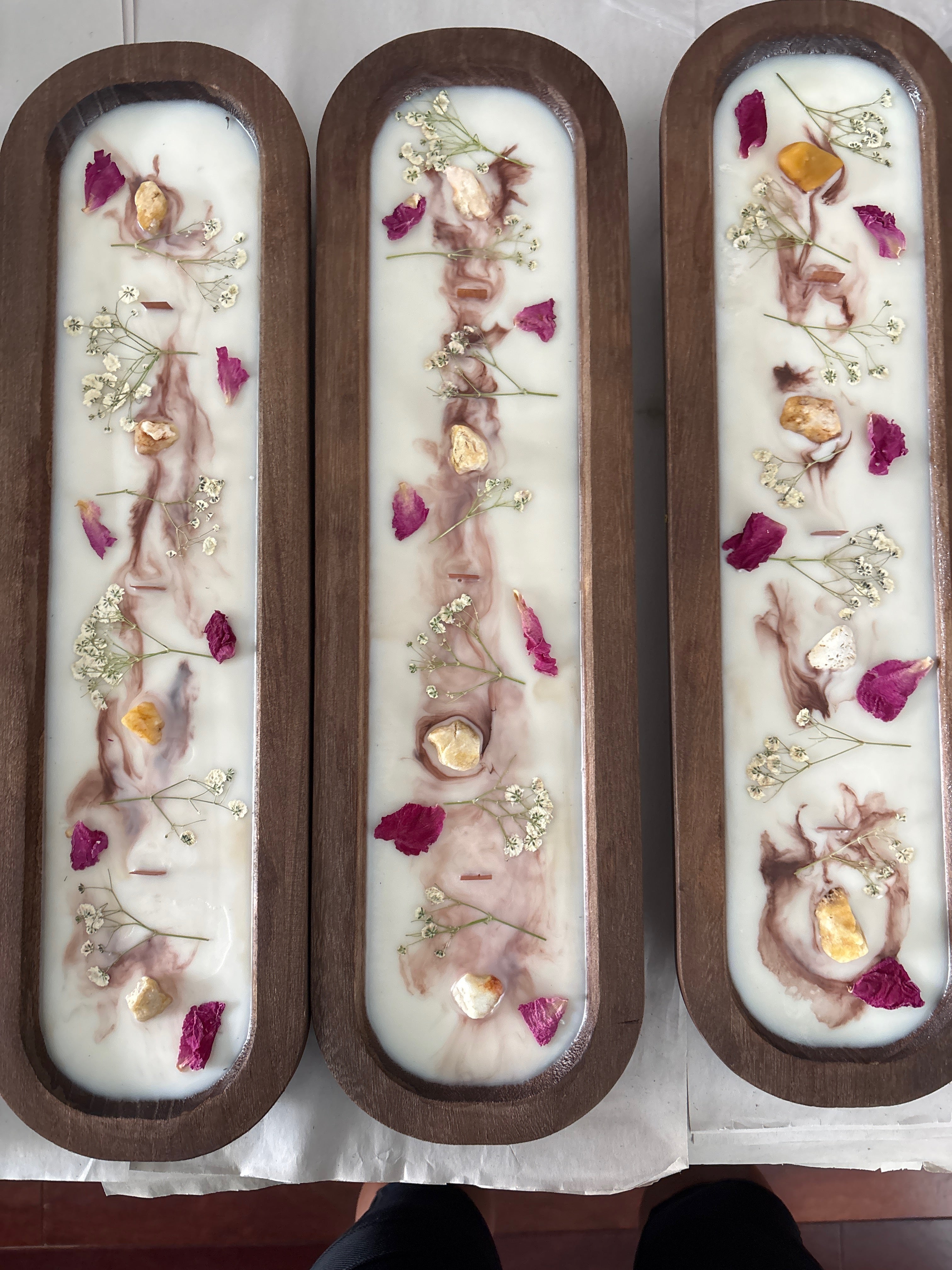 Three long rectangular wooden CANDLES with white wax bases, embedded with dried flowers, petals, and small gold leaf pieces, displayed on a white cloth.