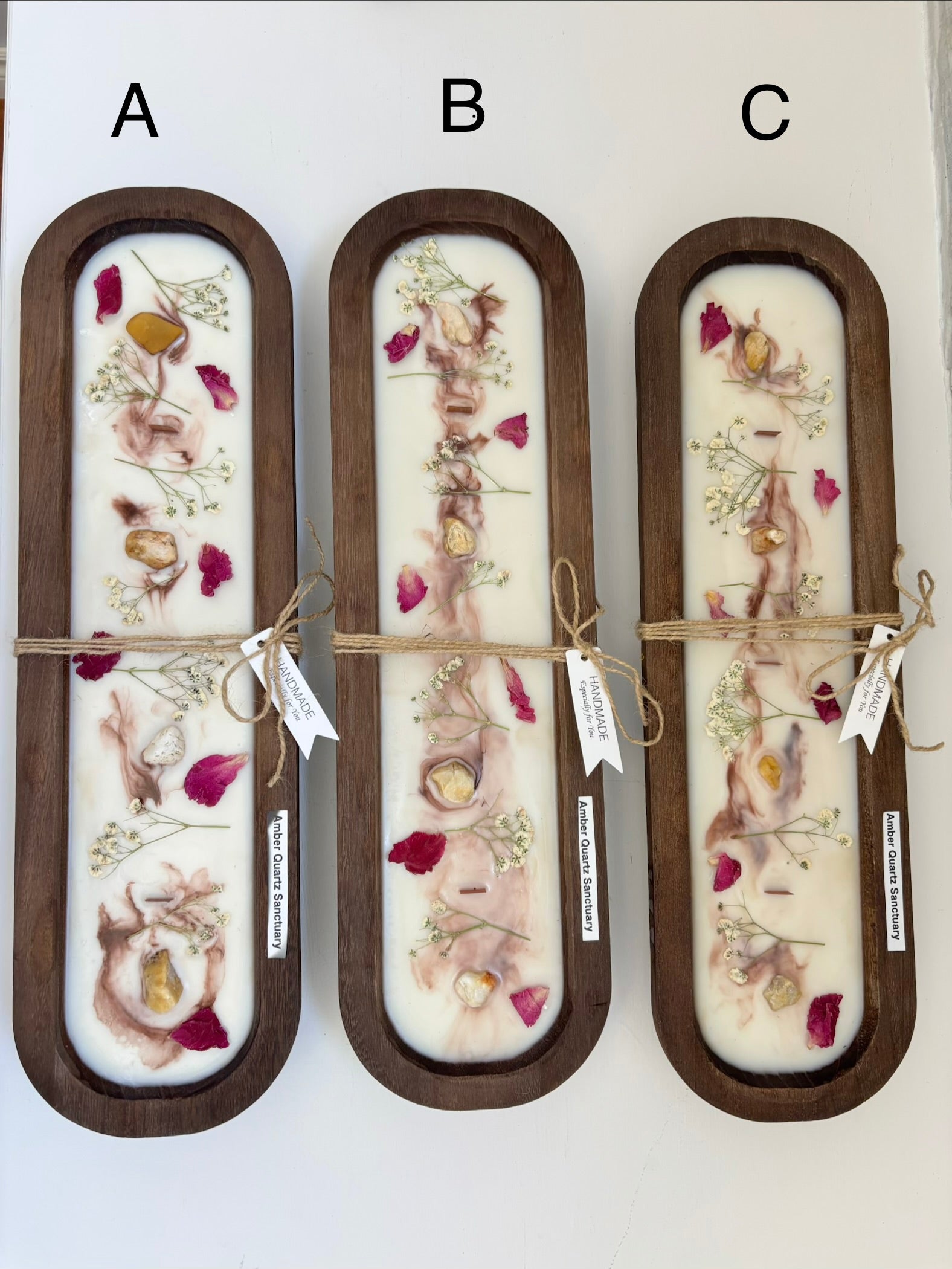 Three long oval wooden trays labeled A, B, and C, each containing a pressed floral arrangement in white resin with pink and red petals, tied with twine and a small paper tag, on a white background.