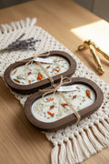 Two oval wooden soap dishes containing white wax candles with dried flowers and botanicals, tied with twine and a small white tag reading HANDMADE.