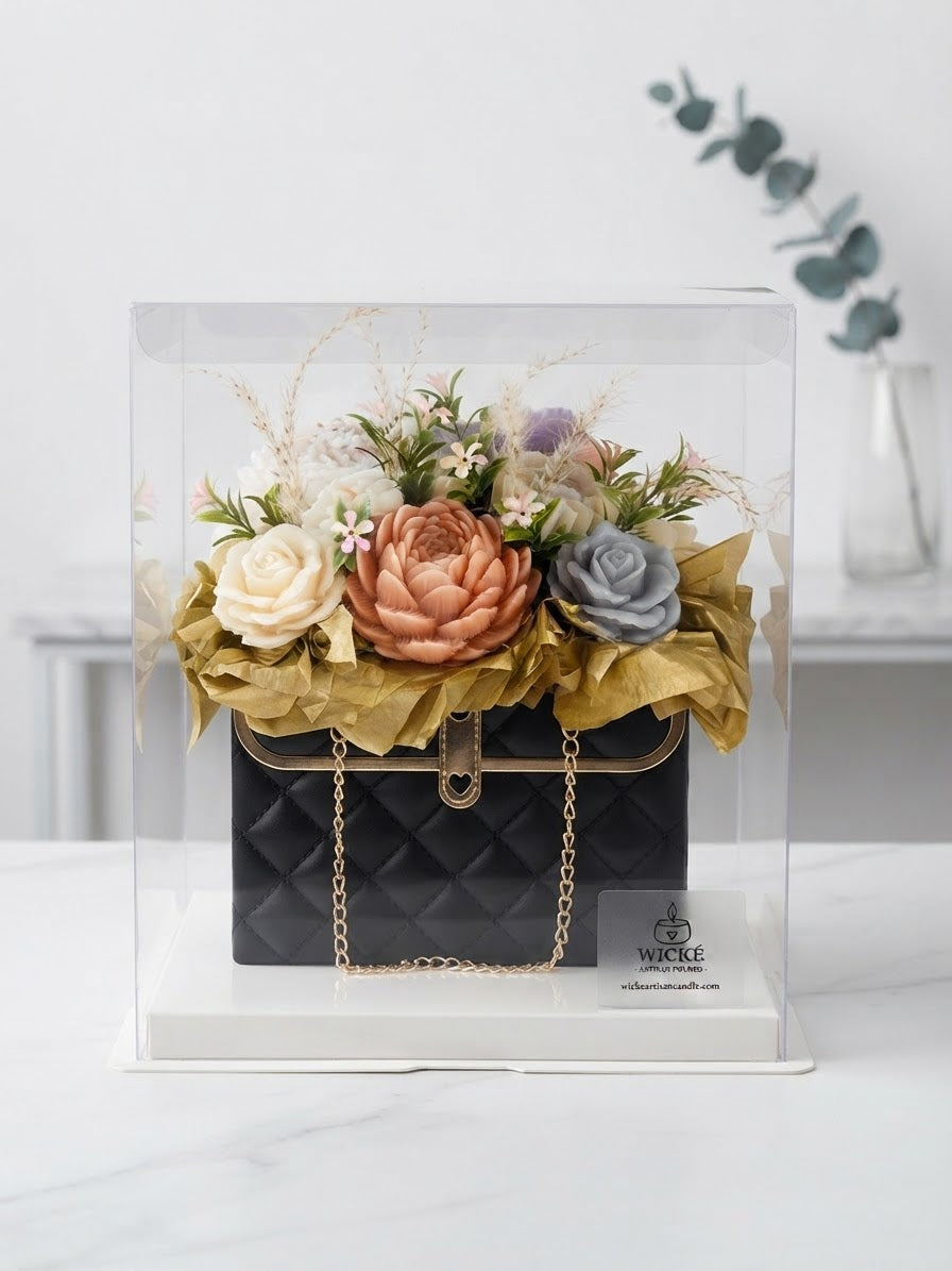 A luxury floral arrangement designed to look like a black quilted handbag with gold chain details, displayed inside a clear acrylic display box. The bag is topped with a bouquet of artificial flowers in peach, cream, and gray, wrapped in gold tissue paper.