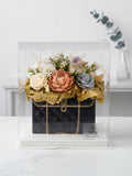 A luxury floral arrangement designed to look like a black quilted handbag with gold chain details, displayed inside a clear acrylic display box. The bag is topped with a bouquet of artificial flowers in peach, cream, and gray, wrapped in gold tissue paper.