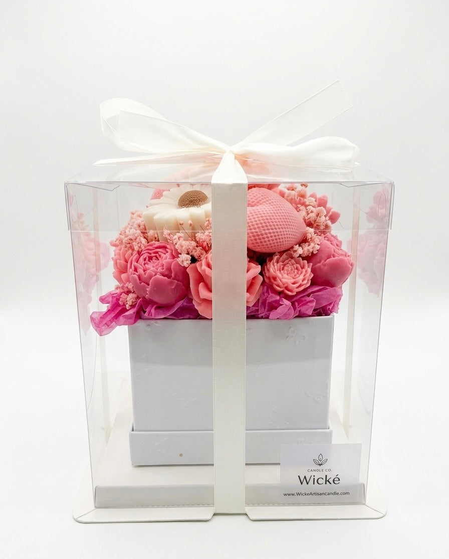Pink floral arrangement in a clear box with a white bow, branded 'Wicke'.
