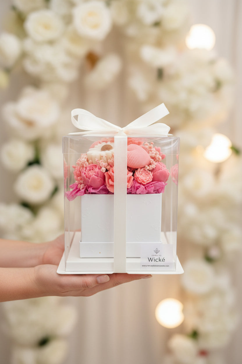 Pink floral arrangement in a clear box with a white bow, branded 'Wicke'.