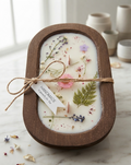 An oval dark wood dough bowl candle filled with white wax, featuring wooden wicks and decorated with embedded pressed flowers and greenery, tied with twine and a "Handmade" tag