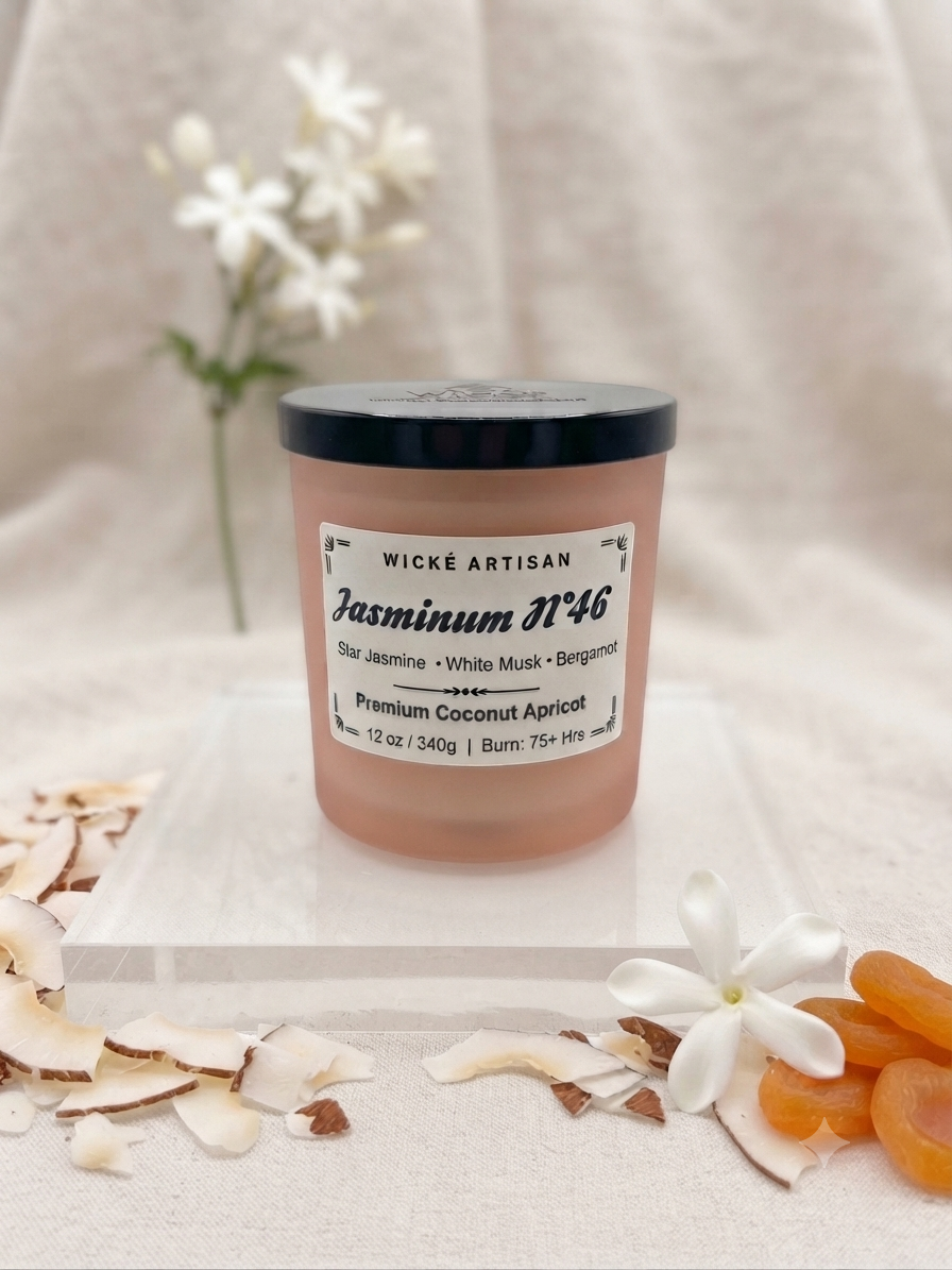 A jar candle labeled 'Wické Artisan Jasminum N°46' sits on a clear acrylic block, surrounded by white flowers, coconut shells, and dried apricots on a textured beige fabric background.