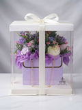 A clear acrylic gift box with a white lid and white ribbon, containing a purple floral arrangement with faux flowers, a lavender box, and gold chain accents. The box is photographed on a light gray studio background.