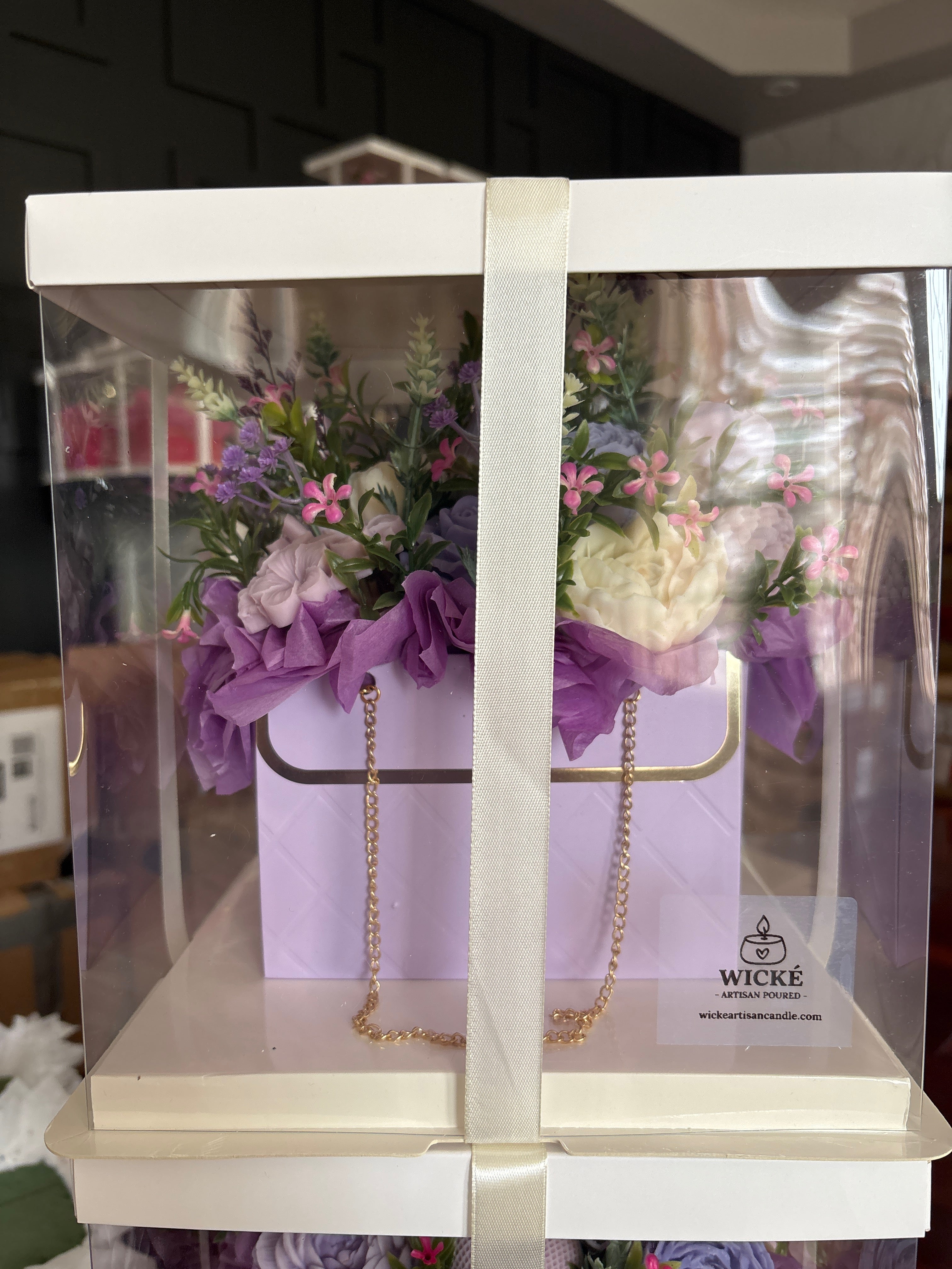 A square floral gift box with a clear plastic outer cover, white interior base, and a white ribbon running vertically down the center. Inside is a lavender quilted flower box holding purple and white flowers with green foliage and pink blossoms.