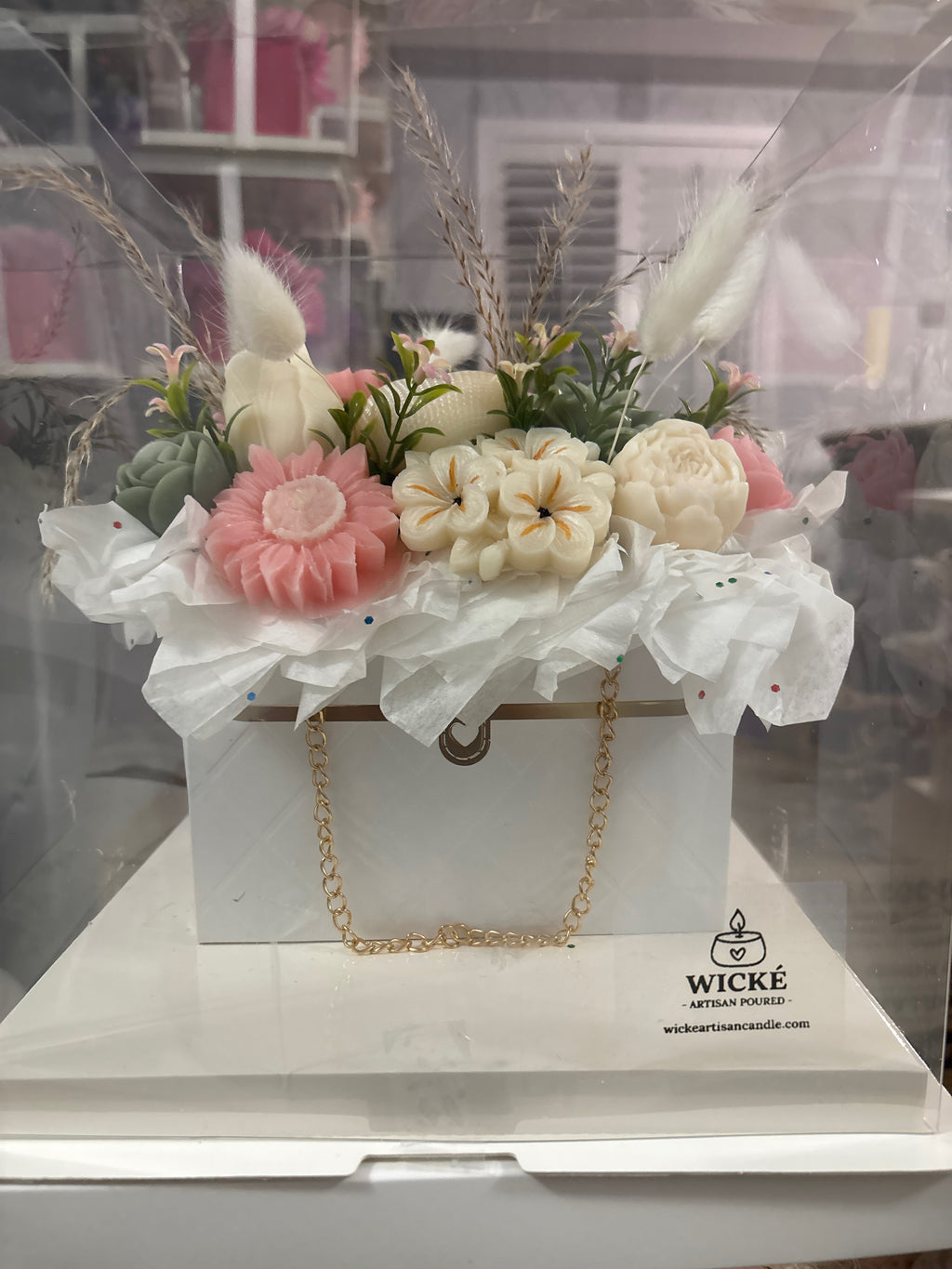 A decorative gift box arrangement by Wick + Artisan Candles, featuring faux flowers in pink, cream, and green, dried grasses, and white tissue paper, presented in a clear box on a white base with a gold chain handle.