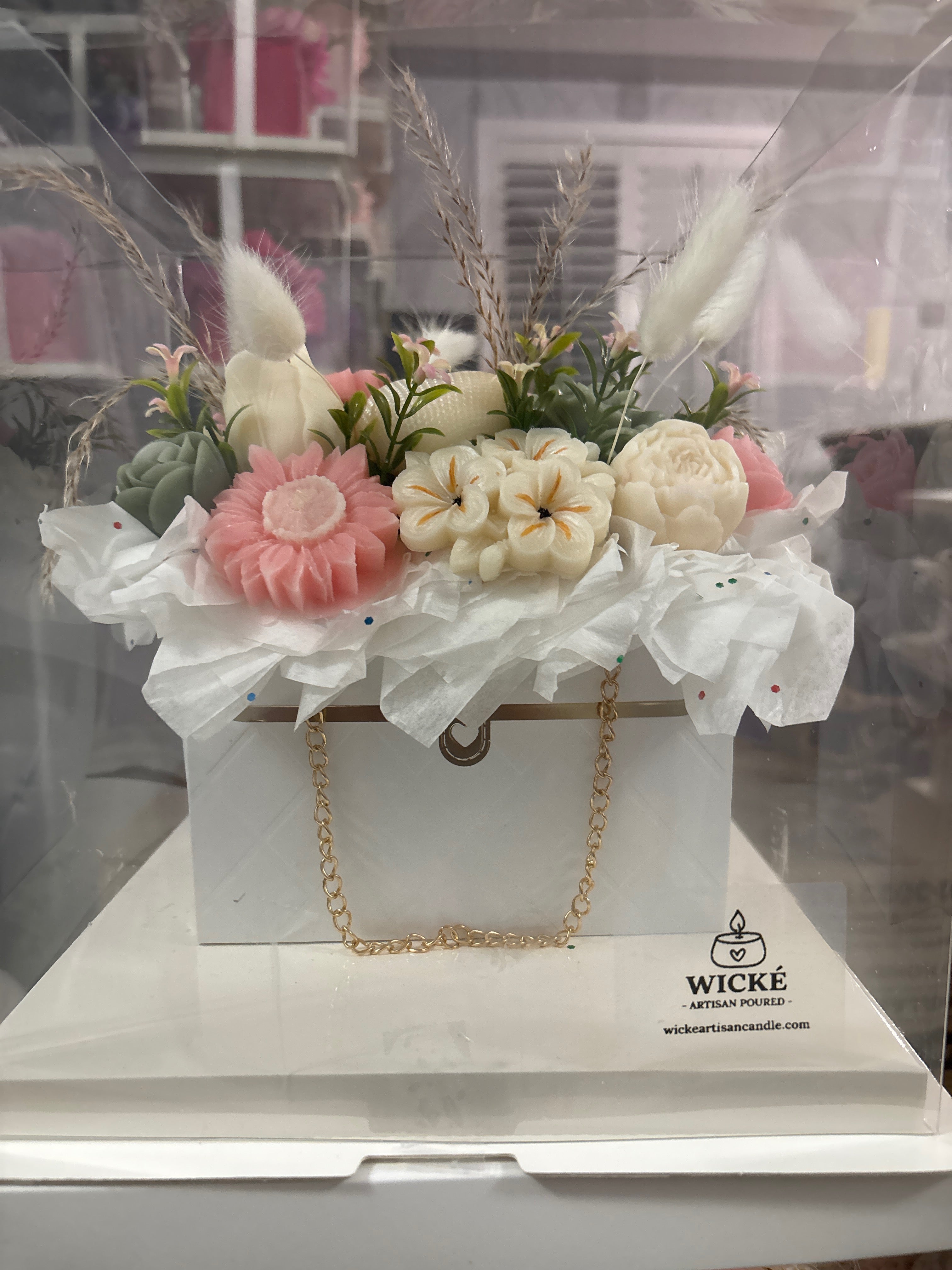 A decorative gift box arrangement by Wick + Artisan Candles, featuring faux flowers in pink, cream, and green, dried grasses, and white tissue paper, presented in a clear box on a white base with a gold chain handle.