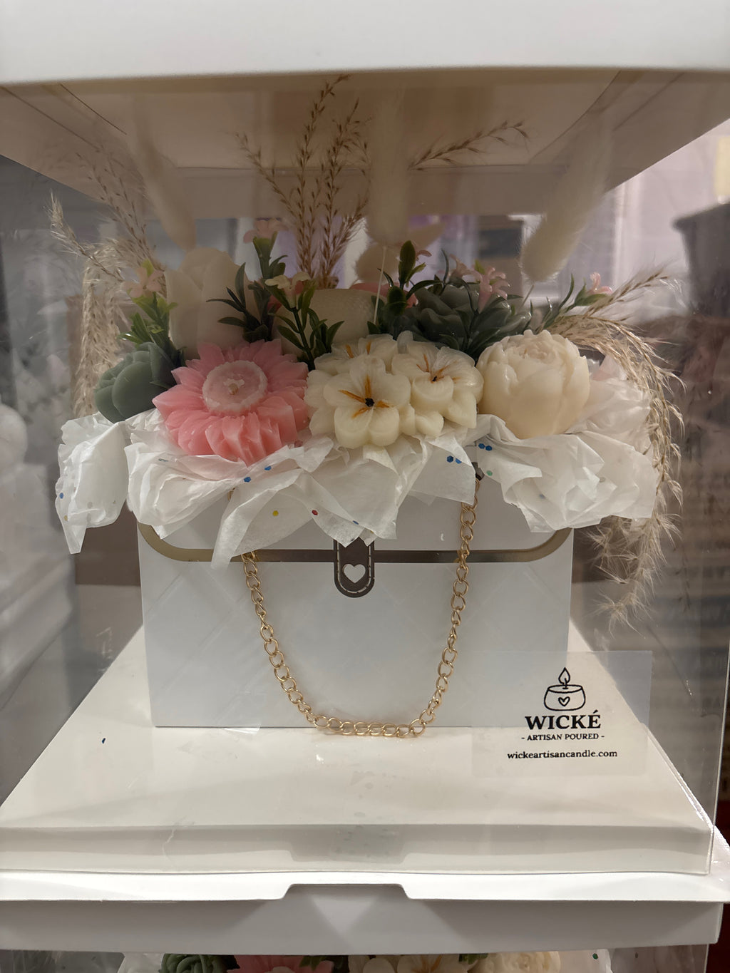 A decorative white gift box with a gold clasp and chain, filled with faux flowers and dried grasses, displayed on a white surface. The box has the Wicke Artisan Candle logo printed on the front.