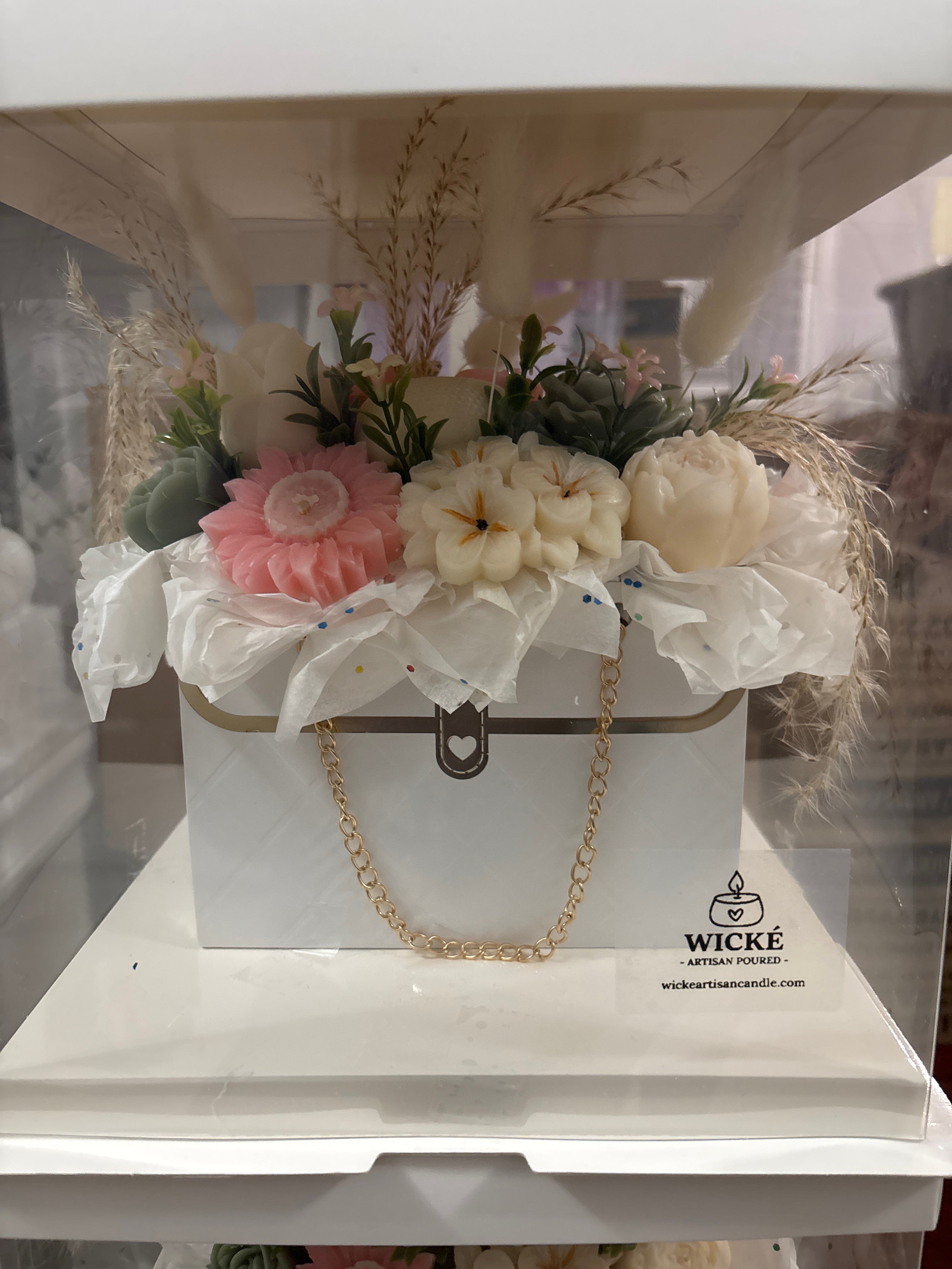 A decorative white gift box with a gold clasp and chain, filled with faux flowers and dried grasses, displayed on a white surface. The box has the Wicke Artisan Candle logo printed on the front.