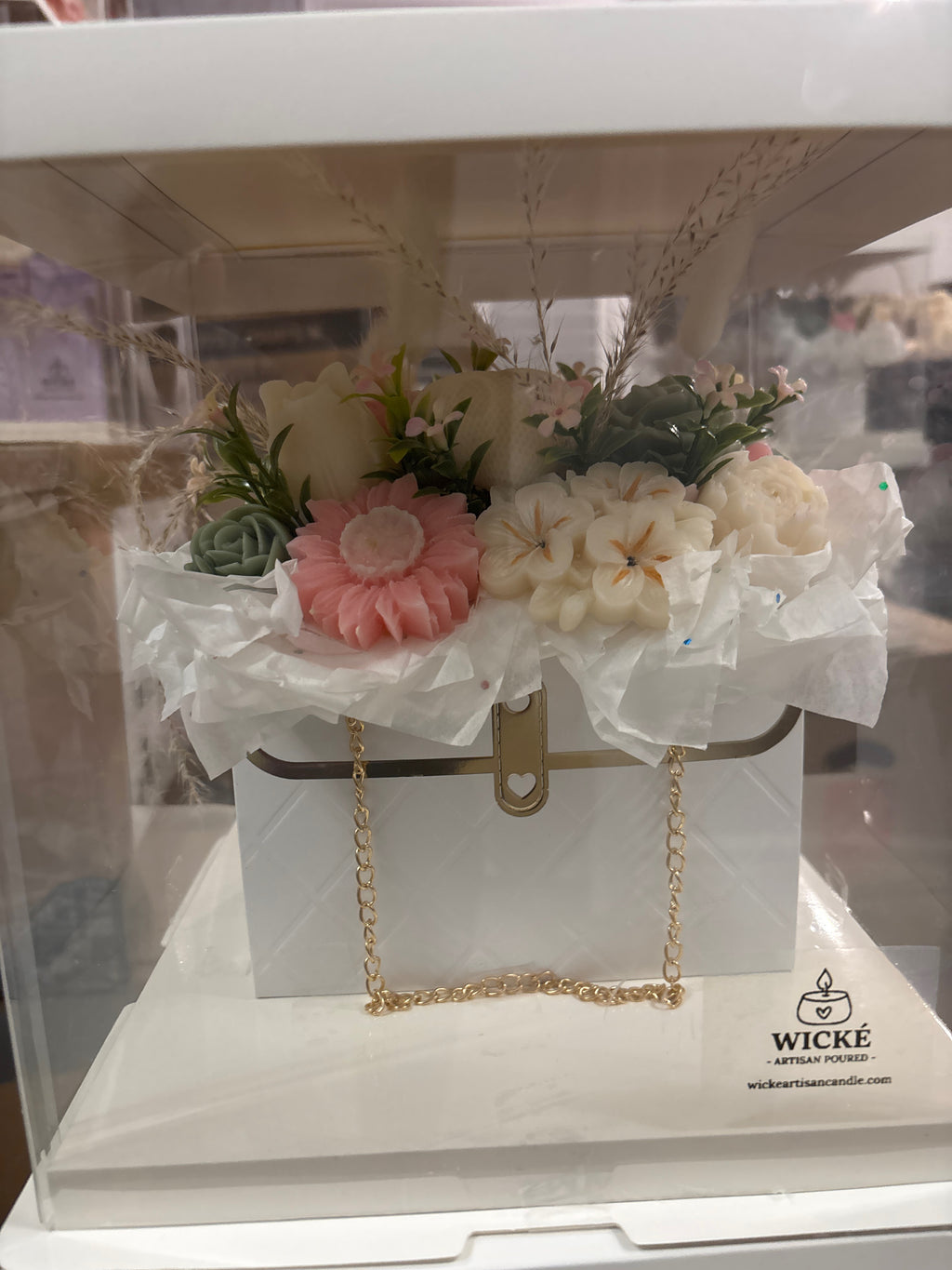 A decorative white gift box with a gold clasp and chain handle, filled with faux flowers and greenery, displayed inside a clear acrylic case. The box has the Wicke Candle Company logo printed on the bottom right.
