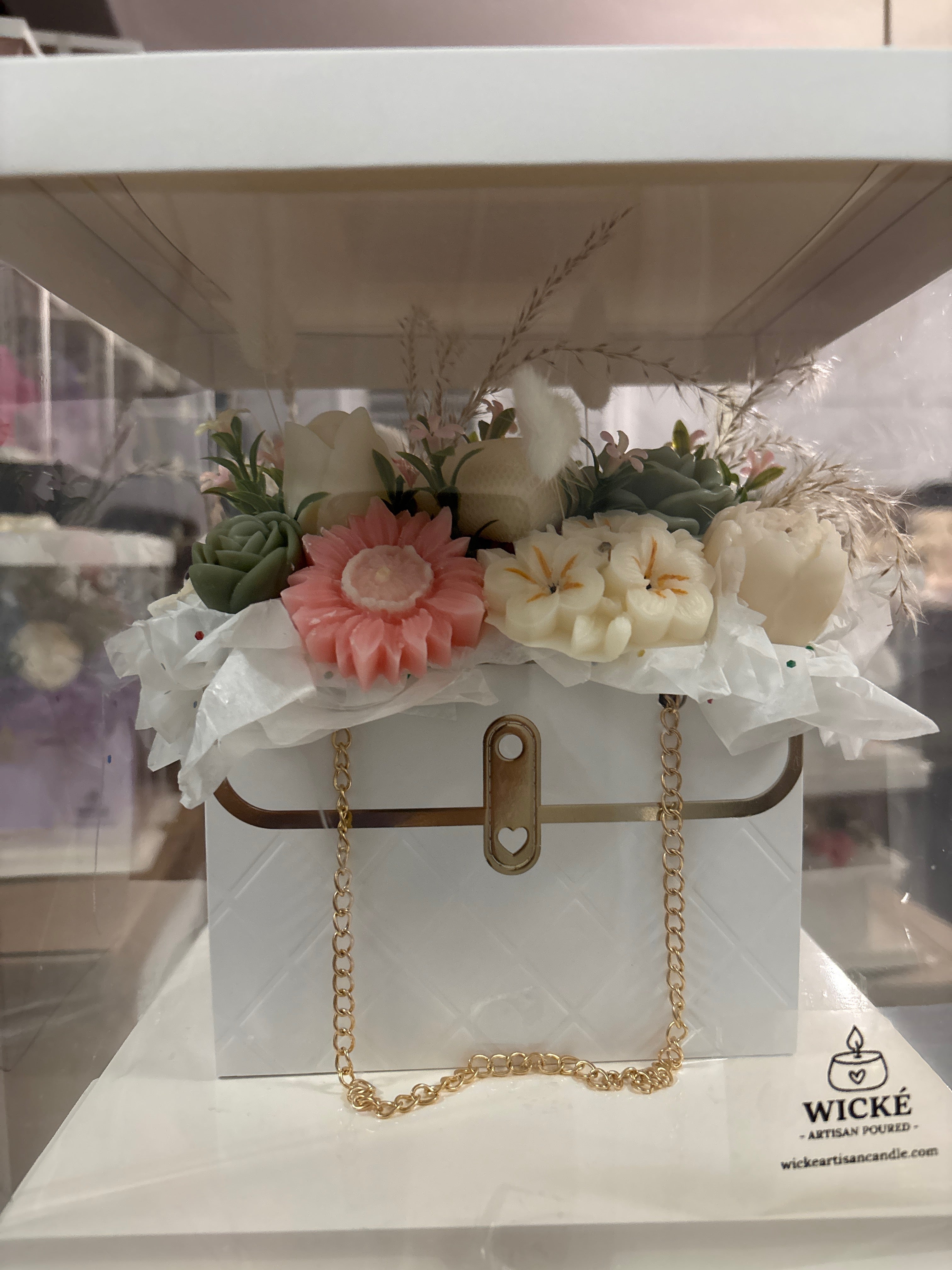 A white quilted decorative gift box with a gold clasp and chain, styled with faux flowers and greenery on white tissue paper. The box sits inside a clear display case, with the WICKÉ Artisan Candle logo visible in the lower right corner.