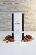 A detailed, single-focused lifestyle product photograph of the Signature Pearl Wax in the Amber scent. A premium black WICKÉ candle box, featuring a bold gold 'W' logo, is isolated and centered on a clean white countertop. It is elegantly surrounded by natural elements like rich, raw amber resin pieces and fragrant sandalwood sticks. Small, loose pearl wax granules are scattered at the base, and soft, golden light highlights the luxurious textures against a clean stone wall background.