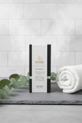 A premium black WICKÉ candle box with a gold "W" logo is centered on a light gray slate surface. It is surrounded by fresh, vibrant eucalyptus branches and a rolled white luxury spa towel. Soft steam gently rises in the background against a minimalist tiled wall, creating an inviting, high-end spa atmosphere.