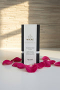 A single black-and-white WICKÉ Pearl Wax Candle box showcasing a clean label is centered on a polished white countertop. It is elegantly framed by lush, dewy English rose petals and a few fresh, deep pink rose blooms. Soft, warm morning sunlight filters in, creating a luxurious and inviting romantic atmosphere against a softly blurred stone wall background.
