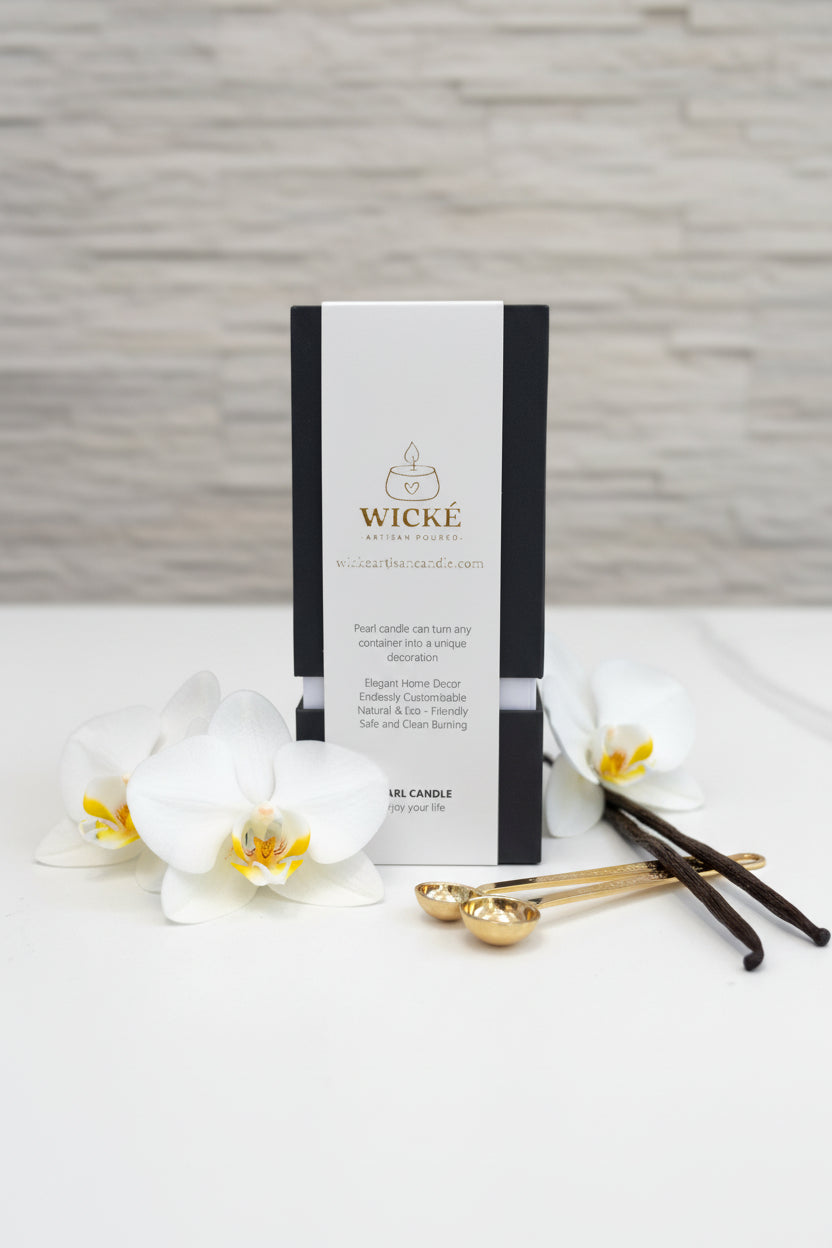An isolated, high-resolution product shot of a single Wické Vanilla Pearl Wax Candle box on a polished white countertop. It is elegantly surrounded by raw vanilla bean pods, delicate blooming white vanilla orchid flowers, and artisanal gold measuring spoons, visually communicating the luxurious fragrance to the viewer against a softened stone wall background.