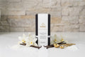 An isolated, high-resolution product shot of a single Wické Vanilla Pearl Wax Candle box on a polished white countertop. It is elegantly surrounded by raw vanilla bean pods, delicate blooming white vanilla orchid flowers, and artisanal gold measuring spoons, visually communicating the luxurious fragrance to the viewer against a softened stone wall background.