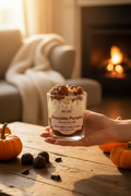 A WICKÉ 'Chocolate Pumpkin' scented candle in a clear glass jar, filled with layers of cream-colored wax, white flower petals, and topped with decorative wax pumpkins and chocolate pieces, set against a plain white background.