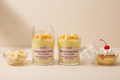 A WICKÉ Banana Crème Glacée aroma candle in a clear glass jar with a clear lid, filled with pale yellow wax and topped with three small yellow banana-shaped wax candles with white drizzle, on a white background.