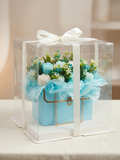 Handcrafted light blue quilted purse candle overflowing with a matching ocean blue and white botanical floral arrangement, showcased inside a premium clear acrylic display box wrapped in a white ribbon.