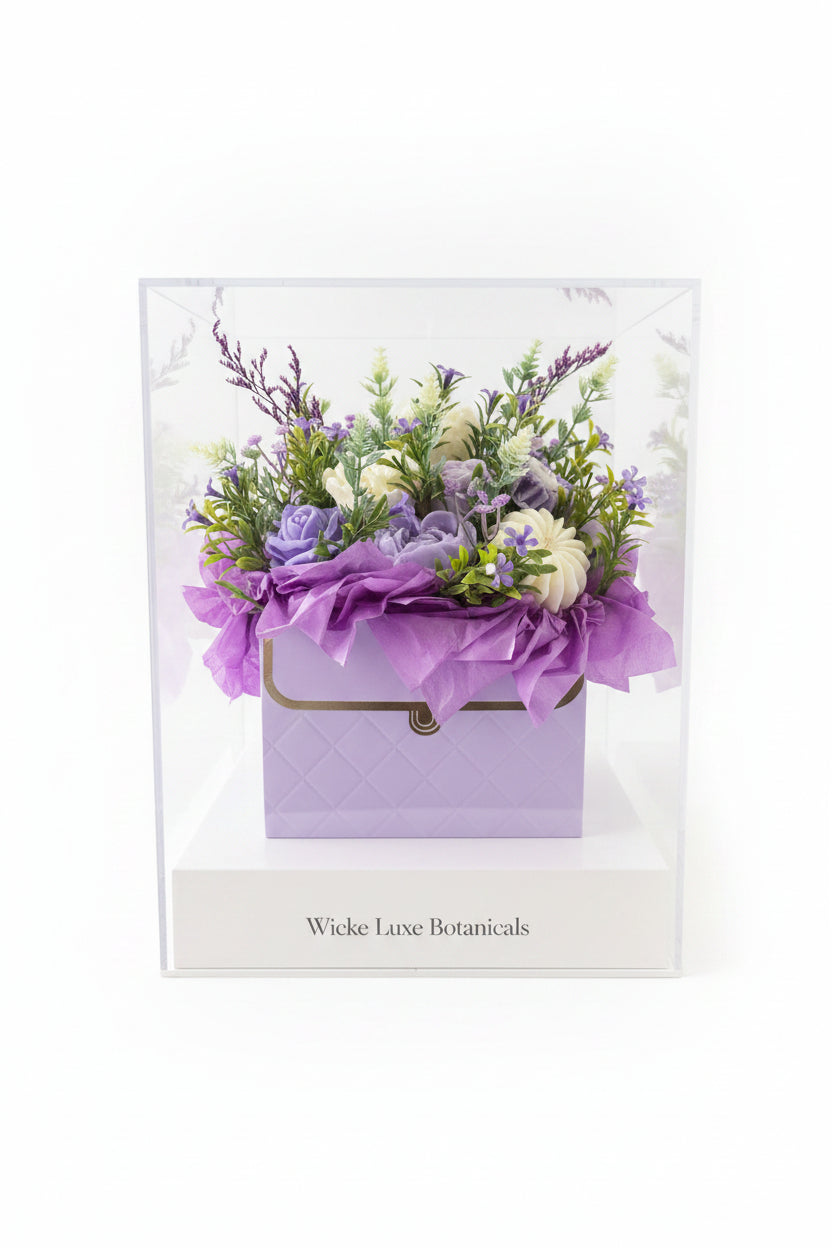 A purple faux flower arrangement in a lavender gift box with purple tissue paper and a gold chain, displayed on a table in a shop setting.