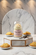 A WICKÉ Butter Cookies aroma candle in a clear glass jar with a clear plastic dome lid. The candle features a layered design with cream-colored frosting swirls, a yellow cracker cookie candle piece on top with small colorful sprinkles, and a pale yellow wax base.
