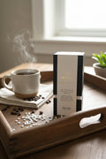 A compelling product photograph of the WICKÉ Coffee Pearl Wax box resting on a vintage wooden breakfast tray. Behind it, a steaming, artisanal ceramic mug of dark coffee sits on a linen napkin. Scattered across the tray are loose, glossy whole coffee beans and pristine white wax pearls. Soft morning sunlight streams from a window, creating a warm, cozy atmosphere that promises a perfectly energized start to the day.