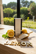 A premium product shot featuring the WICKÉ LEMON Pearl Wax box centered on a textured travertine stone vanity in a sunlit bathroom. Surrounding the box are freshly sliced Sicilian lemons with juice droplets, a sprig of fresh mint, and loose white wax pearls in a polished brass scoop. In the background, out of focus, is a large window with a view of a lush Mediterranean garden, conveying maximum freshness and natural energy. Large, bold brass letters spelling LEMON are arranged on the vanity surface below th