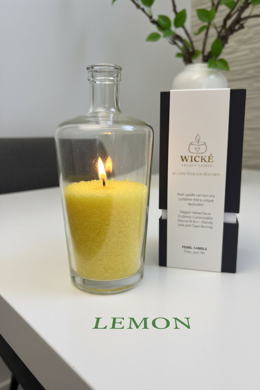 A dramatic close-up focusing on sustainability and creative reuse. A sleek, empty designer gin bottle (like a classic yellow or clear one) has been repurposed, filled with WICKÉ LEMON pearl wax, and is brightly burning. The LEMON box packaging stands prominently behind the vessel. The scene is set on a modern white concrete dining table. Printed in an elegant, eco-friendly green font on the table surface is the word LEMON, linking the customizable experience to natural luxury and conscious decor.