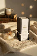 A cozy, inviting close-up photograph conveying comfort and luxury. A single WICKÉ SANDALWOOD box rests on a beautifully textured, chunky knit cashmere blanket. Next to the product, a burning pearl candle in an elegant glass bowl casts a soft, warm golden glow across the fabric. A stack of classic books and a ceramic mug are softly blurred in the background. Printed in a delicate, sophisticated gold font on the edge of the blanket is the word, SANDALWOOD, linking the scent to ultimate relaxation and curated 