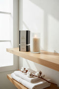 A high-end lifestyle photograph of the WICKÉ White Musk Pearl Wax box resting on a floating shelf made of light, natural oak. Beside it, a minimalist frosted glass cylinder is filled with pristine white wax pearls and a softly burning wick. The background is a sun-drenched, airy bathroom with clean lines, white linen towels, and a single branch of cotton blossoms, evoking a sense of pure serenity and clean luxury.
